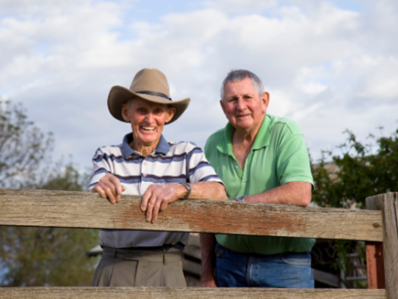 two smiling men, standing outside, leaning against wood fence